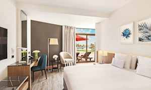 Et hotelværelse med moderne møbler, stor seng, marmorbord og en terrasse med liggestole og udsigt over palmer. (AI-genereret tekst)