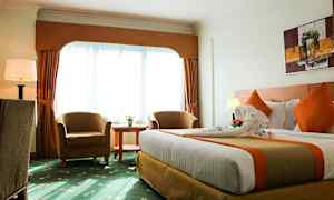 Et hotelværelse med en stor seng, to lænestole, et rundt bord og et stort vindue med gardiner. (AI-genereret tekst)