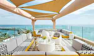 En udendørs terrasse med siddeområder og et skyggetag dækket med stof foran en havudsigt. (AI-genereret tekst)