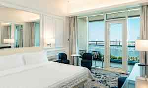 Hotelværelse med redt seng, moderne indretning og balkon med udsigt over havet. (AI-genereret tekst)