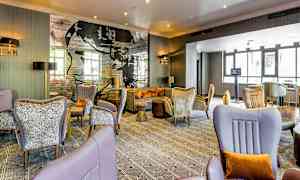 Et moderne hotelrum med komfortable siddemøbler, elegante dekorationer og en vægtapet med tekst og kunst. Store vinduer slipper naturligt lys ind. (AI-genereret tekst)