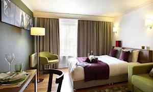 Et hotelværelse med en dobbeltseng med lilla og hvide sengelinned, en grøn stol og et spisebord med service. (AI-genereret tekst)