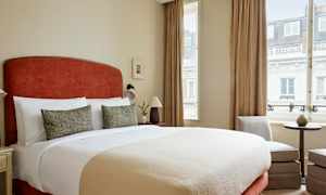 Soveværelse med stor seng, to dekorative puder, lamper ved siderne og vindue med gardiner. (AI-genereret tekst)