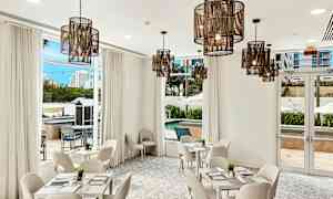 Et lyst og stilfuldt indrettet rum med hvide møbler, store vinduer og hængende lamper. Udsigten viser en pool og omgivende bygninger. (AI-genereret tekst)