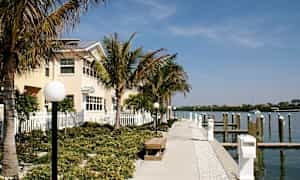 En gangsti med bænke og lamper er omgivet af palmer og fører forbi en bygning til en lystbådehavn. (AI-genereret tekst)