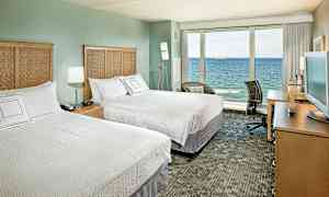 Hotelværelse med to dobbeltsenge, et fjernsyn på et skrivebord og en stor glasvæg med udsigt over havet. (AI-genereret tekst)