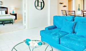 En lys og rummelig interiør med en stue og et soveværelse. I forgrunden ses en blå sofa samt et glasbord med dekorationer. Soveværelset har en seng og en natlampe. (AI-genereret tekst)