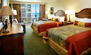 Hotelværelse med to senge med farverige sengetæpper, et skrivebord og en åben balkon med udsigt over havet. (AI-genereret tekst)