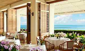 En restaurant med borde dækket med hvide duge og blomster. Baggrunden viser havudsigt med blå himmel. (AI-genereret tekst)