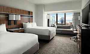 Et hotelværelse med to dobbeltsenge, en siddegruppe, et skrivebord og et fjernsyn, med udsigt over en by gennem vinduet. (AI-genereret tekst)