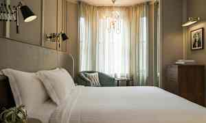 Et elegant soveværelse med en stor seng dækket med hvide lagner, en grøn lænestol og bløde gardiner, der slipper lys ind. (AI-genereret tekst)