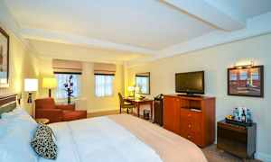 Et hotelværelse med en stor seng, en lænestol, et skrivebord og et fjernsyn på en kommode. (AI-genereret tekst)