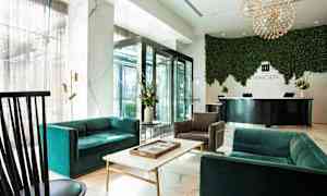 Et moderne hotellobbyområde med sofaer, lænestole og et bord i forgrunden. En reception med skilt ses i baggrunden med en grøn plantevæg. (AI-genereret tekst)