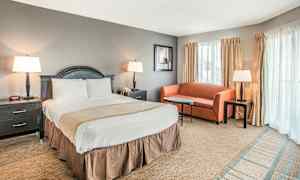 Et hotelværelse med en dobbeltseng, et natbord på hver side, en lille sofa, sofabord og tre lamper. Vinduer med gardiner kan ses i baggrunden. (AI-genereret tekst)