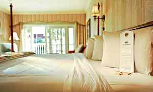 Seng på et hotelværelse med hvide lagner og puder samt tilhørende indretning og lysindfald fra en balkondør. (AI-genereret tekst)