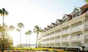 Et hvidt hotel med flere etager og indgange med verandaer, omgivet af palmer og en græsplæne med blomster. (AI-genereret tekst)