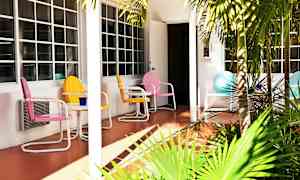 Farverige metalstole og et bord står på en terrasse foran et hus med store vinduer. Foran terrassen er grønne palmer. (AI-genereret tekst)