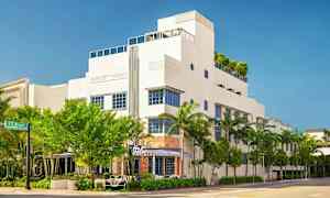 En hvid bygning i moderne stil med blå vinduesrammer, omgivet af træer og palmer. (AI-genereret tekst)