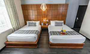 Billede af et hotelværelse med to enkeltsenge med hvidt sengetøj og mønstrede sengetæpper. Rummet har en træpanel på væggen og et natbord med en blomst mellem sengene. (AI-genereret tekst)