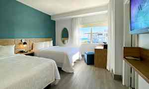 Et moderne hotelværelse med to dobbeltsenge, et skrivebord og et stort vindue, der lader naturligt lys komme ind. (AI-genereret tekst)