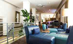 Billede af en hotellobby med blå siddepladser, grønne planter og en bar i baggrunden. (AI-genereret tekst)