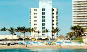 Et hotel med palmer foran, ved siden af stranden med parasoller og mennesker på sanden. (AI-genereret tekst)