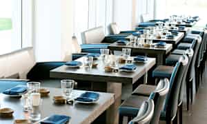 Et elegant restaurantområde med dækkede borde, blå servietter og glas. Lyse vinduer i baggrunden. (AI-genereret tekst)