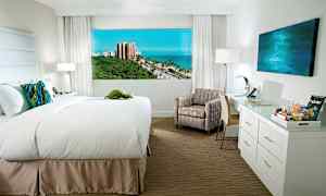 Hotelværelse med en bred seng, stol, skrivebord og udsigt over havet og grønne områder udenfor. (AI-genereret tekst)