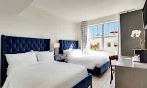 Et hotelværelse med to senge med blå sengegavle, et natbord med lampe mellem sengene og et skrivebord med en blomst og kaffemaskine. (AI-genereret tekst)