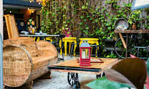 Et udendørs område med en træsofa, et bord med en rød lanterne ovenpå og gule stole i baggrunden. Planter dekorerer omgivelserne. (AI-genereret tekst)