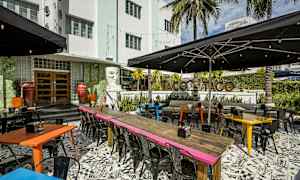En udendørs restaurant med farverige borde og stole i et livligt miljø. Baggrunden viser bygningen og tropiske palmer. (AI-genereret tekst)