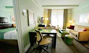 Et hotelværelse med lys indretning, inklusive en seng, skrivebord, sofa og TV. (AI-genereret tekst)