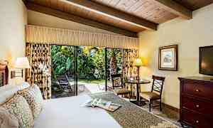 Et hotelværelse med en stor seng, et bord med stole og en åben skydedør, der fører til et frodigt udendørsområde. (AI-genereret tekst)