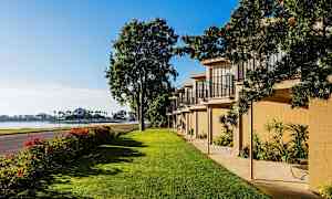 Billedet viser et grønt område med græsplæne, en række bygninger med altaner og en vej ved siden af. I baggrunden ses vandet og palmer. (AI-genereret tekst)