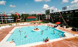 Udendørs swimmingpool med liggestole rundt omkring og folk, der svømmer. I baggrunden ses bygninger med sportstema og en stor dekorativ genstand, der ligner en amerikansk fodbold samt palmer. (AI-genereret tekst)