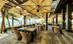 En udendørs restaurant med kurvemøbler og parasoller, beliggende ved stranden med udsigt over havet. (AI-genereret tekst)