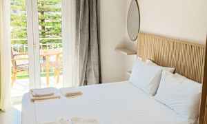 Et lyst og rummeligt soveværelse med en dobbeltseng, hvidt sengetøj og håndklæder. Et stort glasvindue leder ud til en balkon med træmøbler og udsigt over grønne områder. (AI-genereret tekst)