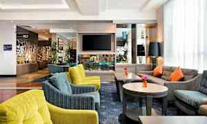 En hotellobby med komfortable siddeområder, flere sofaer og stole i forskellige farver, samt borde med blomster. Et TV er monteret på væggen, og der er dekorative genstande på hylderne. (AI-genereret tekst)