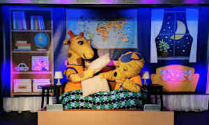 To tegneseriefigurer, en giraf og en bjørn, sidder i en seng med dyne, og scenerekvisitter er synlige i baggrunden. (AI-genereret tekst)