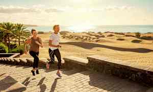 To personer jogger på en stenbelagt gangsti nær sandklitter og palmer ved kysten. (AI-genereret tekst)