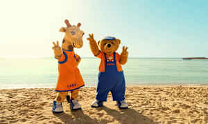 To maskotfigurer med navnene Lollo og Bernie står på en strand foran det klare hav. (AI-genereret tekst)