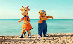 To maskotter, en giraf og en bjørn, står på en strand med havet i baggrunden. (AI-genereret tekst)