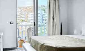 Udsigt fra et hotelværelse med seng og en åben balkondør. Balkon med stole og udsigt over bygninger. (AI-genereret tekst)