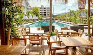 Billede af et hotelområde med liggestole ved siden af en rektangulær pool og træborde i forgrunden. (AI-genereret tekst)