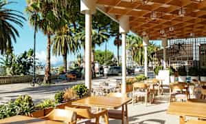 Udendørs servering med træborde og stole, omgivet af palmer og bymiljø. (AI-genereret tekst)