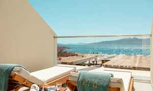 En liggestol med et grønt håndklæde på en balkon med glasrækværk, med udsigt over havet og en strand med parasoller. (AI-genereret tekst)