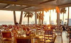 Udendørs spiseområde med borde og stole ved solnedgang, med palmer og havet i baggrunden. (AI-genereret tekst)