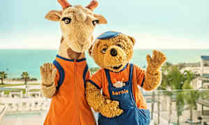 To maskotter, en giraf og en bjørn, står foran havet i baggrunden. (AI-genereret tekst)