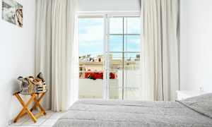 Et lyst soveværelse med en grå opredt seng, en træstol med en sammenrullet taske, gardiner og en altandør, der fører til en udsigt med blomster og bygninger. (AI-genereret tekst)