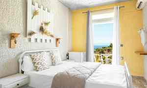 Lyst hotelværelse med seng, dekorerede puder og udsigt til altan med hav og grønt. (AI-genereret tekst)